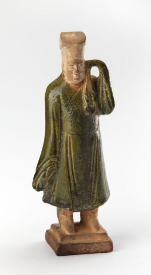  A small figurine depicting a standing person in long, draped robes with elongated, featureless head and raised arm, done in mottled green and brown hues, on a white background. Artist name and title unknown.