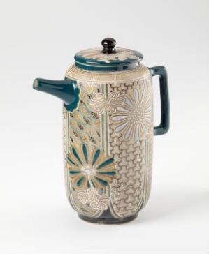  An ornate ceramic coffee pot with a creamy white base, detailed geometric and floral patterns in muted greens, teals, and grays, a teal-colored handle and lid trim, and a simple white spout, all against a light background.