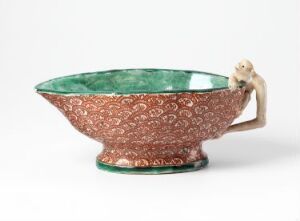  An ornate, wide bowl with a rich reddish-brown exterior featuring white swirling patterns and a sculptural cream-colored monkey as a handle on the side. The interior of the bowl is a striking emerald green. The bowl rests on a plain background, highlighting its intricate design and vibrant contrasting colors.