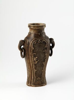  An antiqued bronze vase with a flared rim and tubular body, featuring embossed floral designs and sculpted lion-headed handles, set against a neutral background.