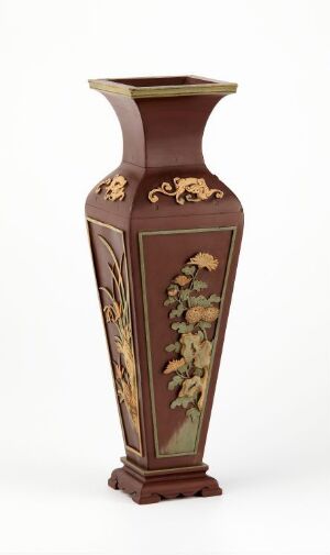  An ornate hexagonal vase in rich brown with a painted floral design in creams, yellows, and greens on the front. The vase has an elegant neck with a lighter color band just below the opening, set against a white background. Artist name and title are unknown.