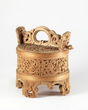 
 An intricately carved light brown wooden object on a stand with floral motifs, featuring a hollow interior, detailed patterns, and a handle on the top.