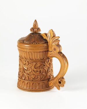  A traditional, decorative beer stein made of a warm golden-brown, wood-like material, featuring intricate foliage carvings, a domed lid, and an ornate handle. The stein is set against a plain white background, highlighting its detailed design and craftsmanship.