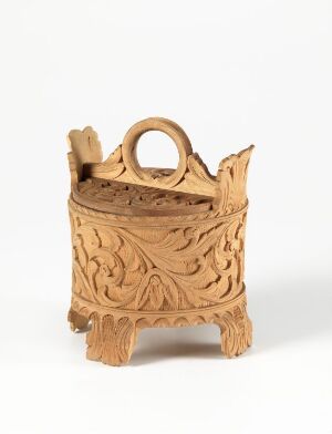  A light to medium brown intricately carved wooden object on a neutral background, resembling a mini decorative basket or container with a looped handle and four feet. Artist name and title unknown.