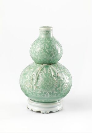  An elegant double-gourd vase with a soft celadon glaze, featuring intricately patterned floral embossing, displayed against a light gray background.