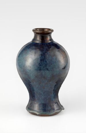  A blue glazed ceramic vase with a narrow neck and rounded body, showcasing shades of deep blue with hints of purple and speckles, creating a textured and luminous effect against a soft white background. Artist name and title are unknown.