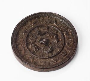  An antique, dark brown, bronze-like metal disc with intricate embossed designs and symbols, featuring a central motif surrounded by concentric circles of what may be text or iconography, against a light background.