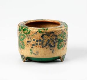  A small hexagonal ceramic pot with a cream beige background and green foliage patterns, featuring leaves and grape-like clusters, with a rich brown glazed rim on top and a green bottom rim.