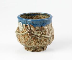 
 A ceramic cup with a blue interior and a textured tan exterior decorated with brown branches and small white flowers with yellow centers. The cup has an organic, rustic shape, and is set against a plain white background. Artist name and title are unknown.