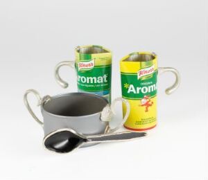  "Tin Mugs" by Hans Stofer, featuring a grey metal mug with a welded tin handle, and two repurposed commercial cans with handles attached, all standing on a light background. The first can is green with a darker green stripe, and the second can is yellow with a green stripe and red text, all evoking a mixture of functionality and art.