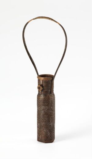  An elongated, cylindrical handwoven object with a round, looped handle at the top, showcasing a rich, earthy brown color and intricate texture, set against a plain white background. Artist name and title are unknown.