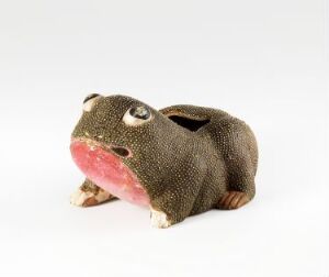  A small figurine of a frog made from a textured, earthy green fabric with speckles, featuring prominent white and black eyes, an open mouth with a gradient pink interior, and stubby beige legs, against a plain white background.