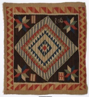  A photograph of a square quilted textile featuring a complex geometric pattern with a central concentric diamond design in blue, yellow, and red, surrounded by red and green triangular motifs and a red and tan striped border, set against a black background with green and red corner accents, and a handwritten inscription "Red".