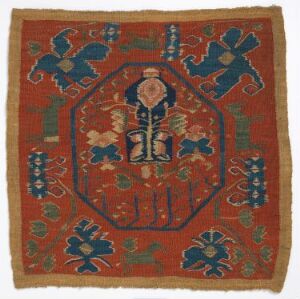  An antique textile, possibly a rug or tapestry, with a deep terracotta background and a central medallion of indigo and light blue, featuring a symmetrical plant-like design in ivory. The piece is bordered by similar geometric floral patterns and displays signs of age. Artist name and title are unknown.