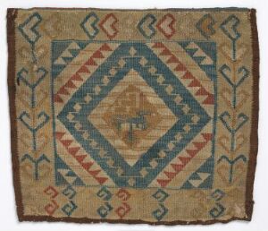  An antique square quilt with a central, diamond-shaped design featuring a bird silhouette surrounded by concentric geometric patterns in earthy tones of tan, rust, and dark blue, with stylized heart motifs and additional geometric shapes in the borders.