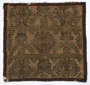  A square, woven textile featuring a symmetrical pattern with shades of beige, blue, green, and dark brown, showcasing a possibly traditional design, with some edges appearing frayed and worn.