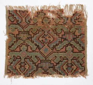  A fragment of an ornate woven textile, showcasing a geometric pattern in shades of brown, cream, green, and faded rust, with fringed edges, displayed against a light background.