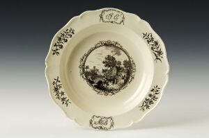  An antique decorative plate with a creamy off-white base and detailed black designs featuring floral patterns around the rim and a central pastoral scene with a bridge, trees, and figures, suggesting a peaceful landscape.