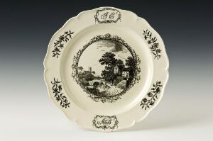  Vintage white ceramic plate with a central monochromatic pastoral scene and decorative floral patterns around the rim, all in a sepia or dark gray tone, against an off-white background.