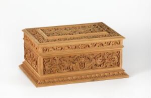  An intricately carved wooden chest with a hinged lid, displaying detailed floral or vine-like motifs, set against a light grey background. The chest has a warm ivory hue with visible wood grain.