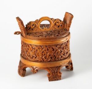  An ornately carved wooden container with a detailed floral motif sits on a plain background. The cylindrical container boasts a domed, intricately carved lid with a handle on the top, all supported by a carved base with four elegant legs.