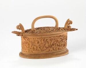  A light brown terracotta casserole with intricate carvings and scrollwork, featuring a domed lid with a central handle, and stylized side handles, set against an off-white background. Artistname and title are unknown.