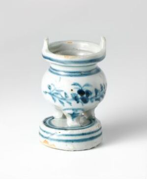  A small white ceramic vase with traditional blue floral patterns and decorative flourishes, featuring a flared neck with scalloped rim and two small handles, displayed against a plain background. The artist and title are unknown.