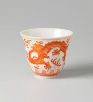  A ceramic cup on a plain background, adorned with a detailed orange dragon design against an off-white surface. The dragon's body wraps around the cup with intricate scales and mane visible in the vibrant glaze.