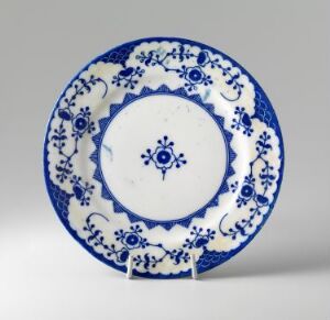  A ceramic plate with a blue and white design featuring a central blue flower-like motif and a highly ornate rim with stylized florals and foliage, presented against a soft grey background.