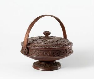  An intricately carved, dark brown wooden container with a pedestal base and an overarching handle, featuring detailed floral and geometric patterns. Artist name and title remain unknown.