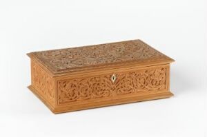  A light brown rectangular wooden box with intricate carvings, set against a simple light background, featuring floral and geometric patterns, and a visible keyhole on the front.