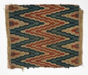  Aged textile with a geometric wave-like zigzag pattern in earthy colors of rust, mustard, dark teal, and navy blue. The pattern creates a three-dimensional optical illusion of movement and the textile has frayed edges, indicating it may be antique.