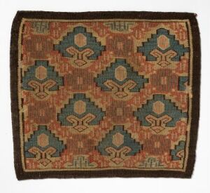  A square textile with a symmetrical geometric pattern featuring teal, beige, and navy blue shapes on a terracotta background, bordered by a dark band, likely a rug or tapestry.