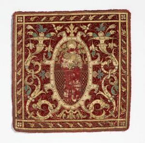  A square textile piece with a rich crimson red base color, featuring an opulent beige border decorated with dark red and green floral and acanthus leaf motifs, and an intricate oval medallion centered with a heraldic crest in matching colors.