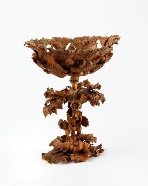  A highly detailed, brown ornate goblet-like object with leaf and floral patterns carved in high relief, set against an off-white background.