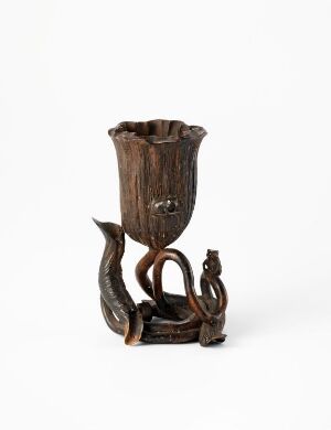  A dark brown, intricately carved wooden cup or chalice with a textured floral-like upper edge and a base resembling intertwining roots or branches, with a serpent-like creature sculpted around it, set against a white background.