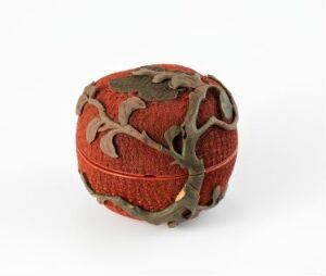  An intricately designed spherical object with a deep red, woven texture core enveloped by a network of dull gray ornamental metalwork, set against a neutral off-white background.