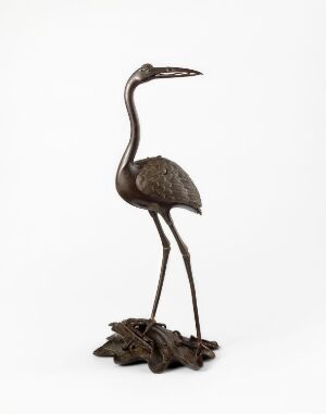  A bronze sculpture of a heron standing on one leg with intricate feather details, set against a white background. The bird's neck is gracefully curved and its beak points slightly upwards. It stands on a base resembling foliage.