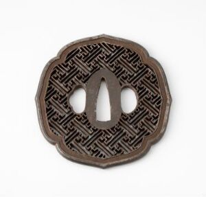  An ancient circular medallion with a scalloped edge and a symmetrical maze-like pattern engraved into a dark brown metal surface, featuring a central elongated opening with a pointed shape.