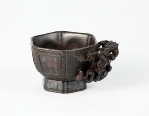  An antique dark brown hexagonal cup with decorative embossed panels and an attached cluster of organic forms on one side, possibly made of metal or carved wood, displayed against a neutral background.