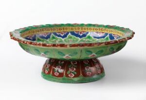  A traditional Thai Bencharong shallow porcelain bowl with a flared, scalloped rim and intricate multi-colored patterns, predominantly green, white, yellow, blue, and red, on a white background. Artist Unknown.