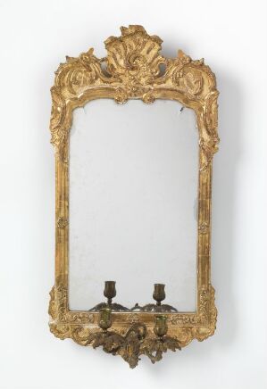  An antique golden gilded mirror with a highly ornate frame featuring a decorative crest at the top, intricate scrolling motifs, and candle sconce holders at the bottom, set against a plain white background.