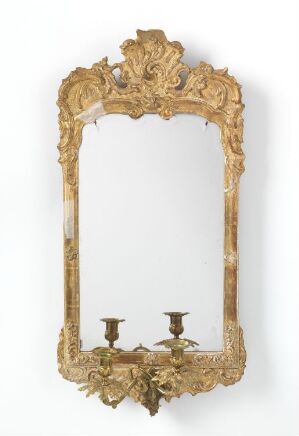  An antique-style gold-framed mirror with intricate floral and acanthus leaf designs throughout, featuring two candle sconces attached to the bottom of the frame, against a plain white background.