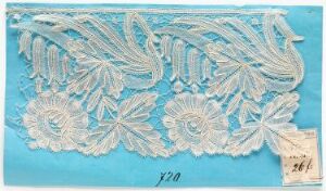  A white lace sample with an elaborate floral and foliage embroidery pattern on a turquoise blue background, accompanied by a small paper label with handwriting.