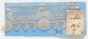  A piece of creamy white lace with an elaborate pattern is mounted on pale blue paper, accompanied by a small beige card with handwritten cursive text. Artist name and title are unknown.