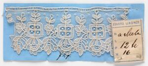  A piece of antique white lace with symmetrical floral and geometric patterns mounted on blue paper, with a small handwritten tag in the upper-right corner.