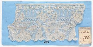  An ornate off-white lace piece with floral and geometric patterns displayed on a light blue background, accompanied by a small handwritten label in the lower right corner.