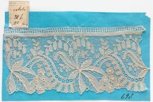  A photograph of an intricate lace design featuring a large central floral pattern surrounded by leaves and smaller flowers, against a light turquoise to sky blue background with a rectangular label in the top left corner.