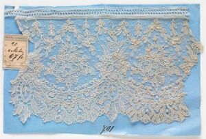  A piece of intricately designed, creamy beige lace with floral and leaf-like patterns is laid on a solid blue background, accompanied by a paper label with handwritten text in the top left corner.