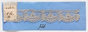  A fragment of white or off-white lace with a repeating floral or foliate pattern is mounted on a medium blue paper background, with an aged paper label in the top left corner, displaying handwritten text.